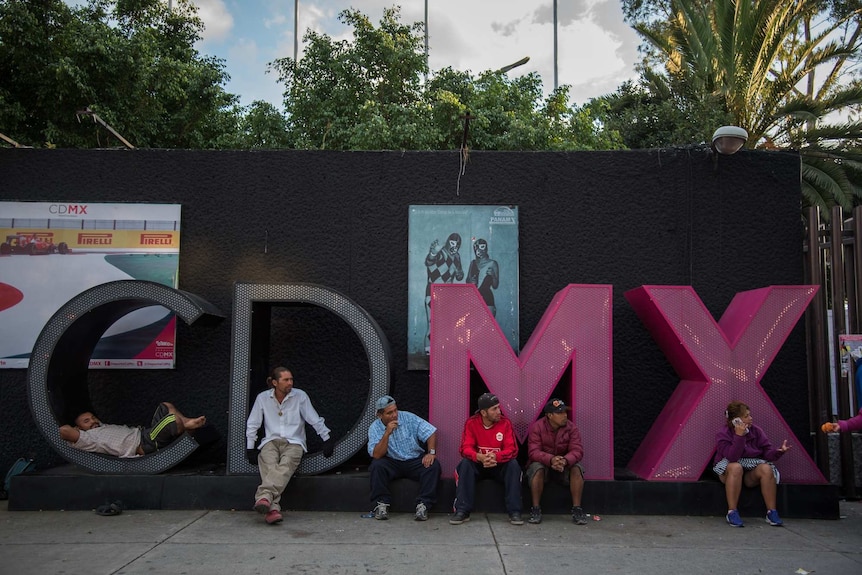 Central American migrants rest and phone home while sitting in big letters spelling CDMX