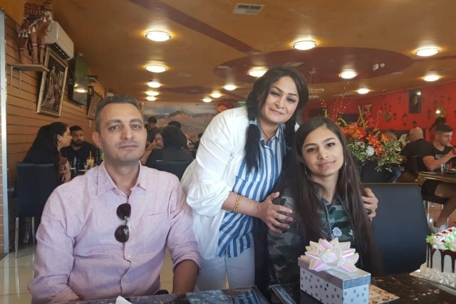 A man sits next to a woman who is affectionately holding the shoulders of a teenage girl, they are sitting in a restaurant