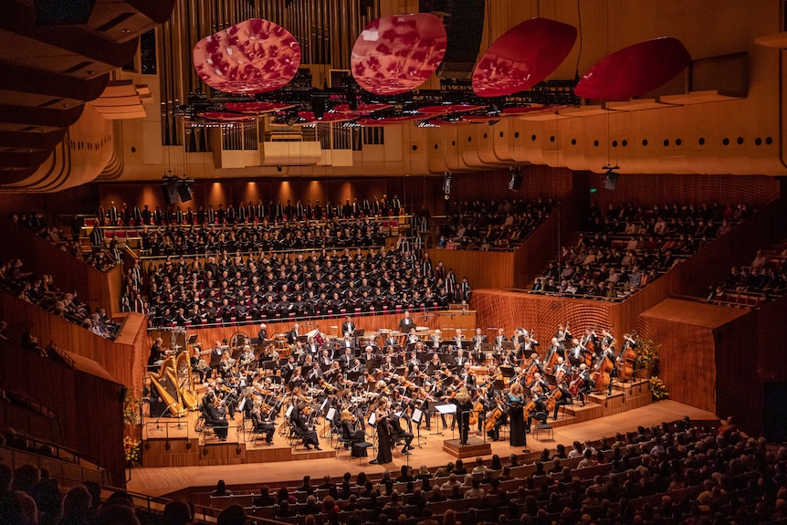 A large orchestra and choir performs on the Sydney Opera House stage. Magenta acoustic reflectors are suspended from ceiling. 