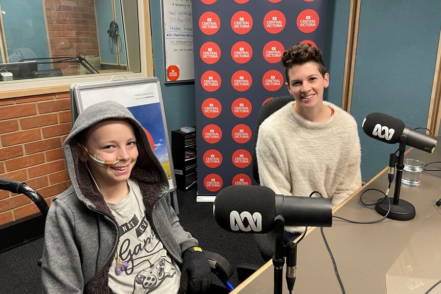 A teenager wearing a hoodie, using a wheelchair, sitting next to his mum in a radio studio.