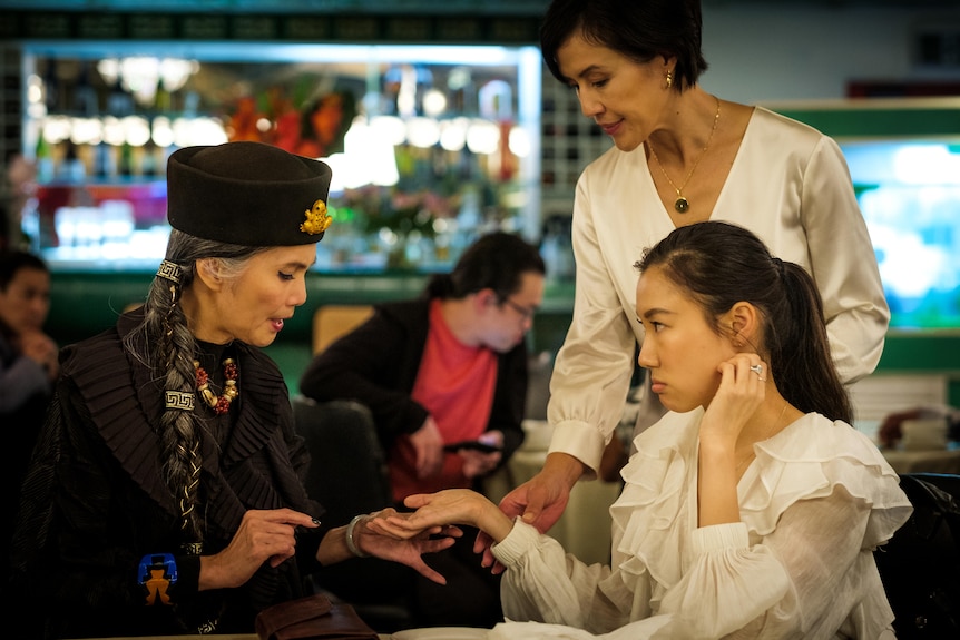 An older woman wearing a black hat with a long grey braid reads the hand of a young woman, whose mother stands above her.