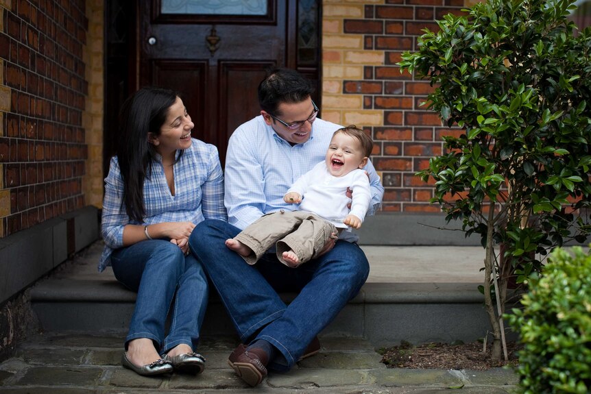 A mum and dad sit on a step in front of their house while holding their giggling son