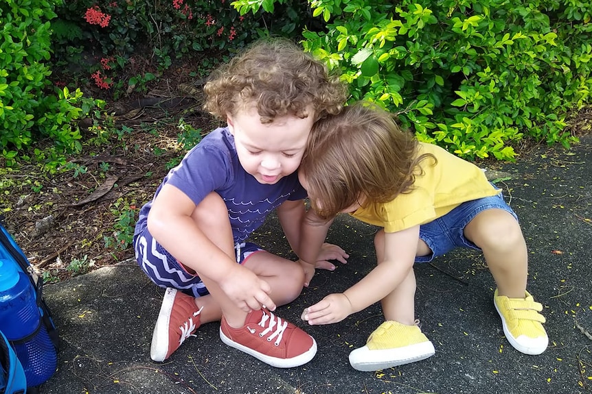 Two toddlers, one with curly brown hair, and one with straight, crouch on the ground playing together.