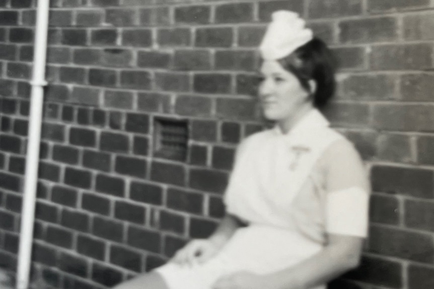 An old black and white photograph of a young woman in a nurse uniform sitting against a brick wall