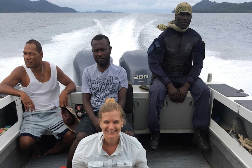 Female sitting on boat floor, three man behind her on ledges of boat, driving through water.