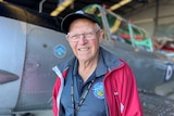 An older man wearing a red jacket and cap stands in front of a vintage plane.