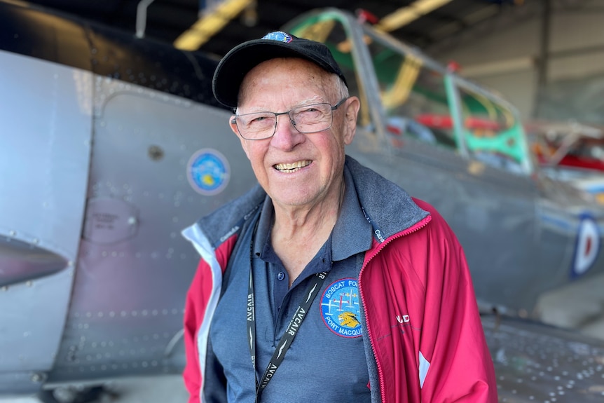 An older man wearing a red jacket and cap stands in front of a vintage plane.