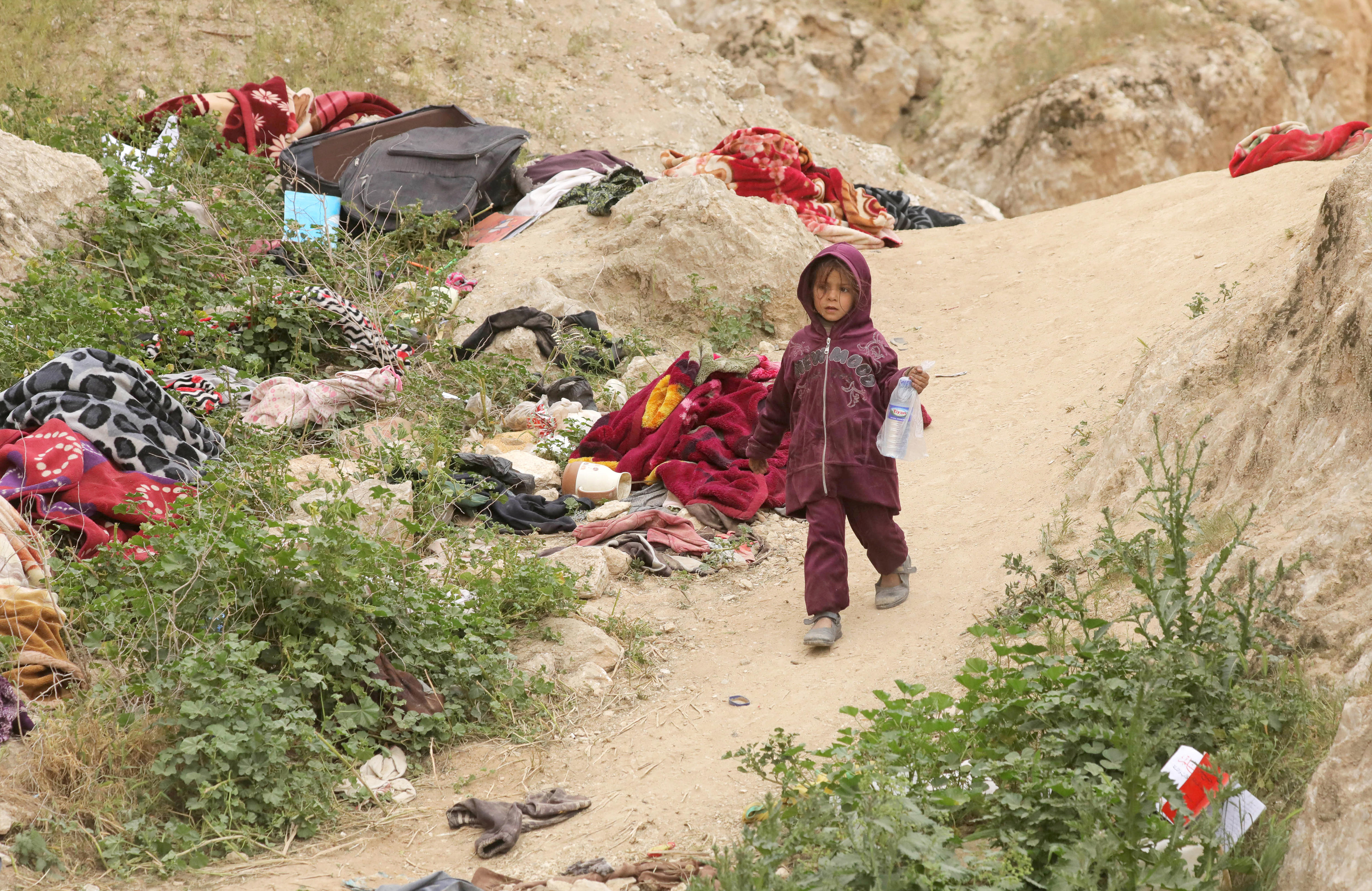 A girl in a hoodie walks among abandoned belongings in a desert landscape.