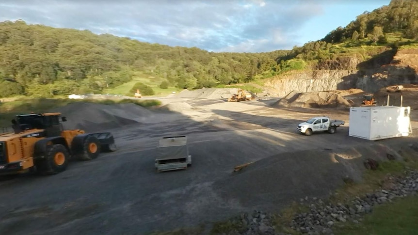A quarry with a tractor and two trucks, green trees in background 