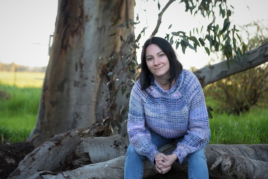 A smiling woman with dark hair, blue and pink sweater, jeans, sits on a cut down tree, grass behind.