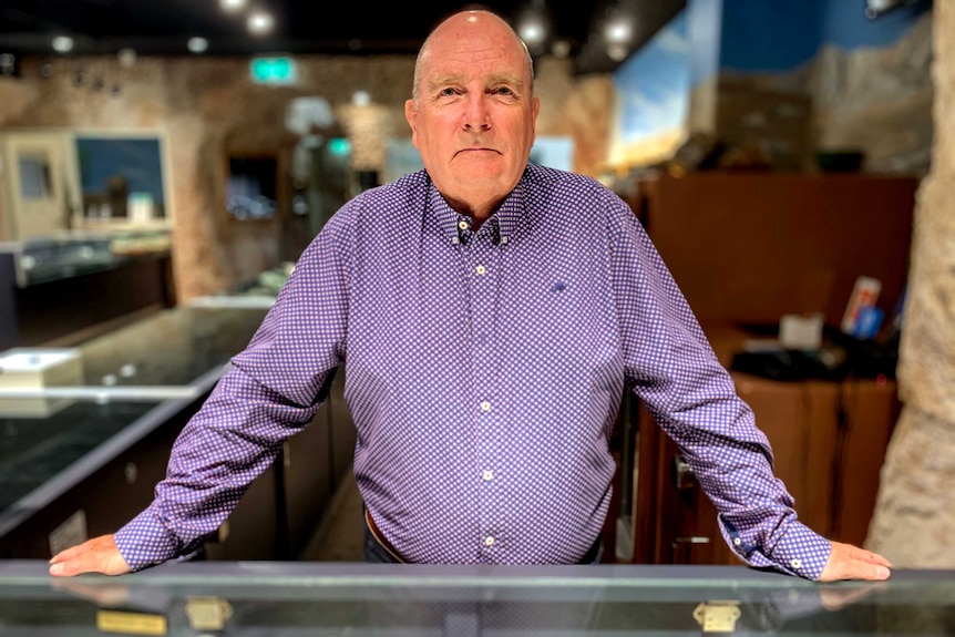 Bald man wearing purple shirt stands with at counter with store in background