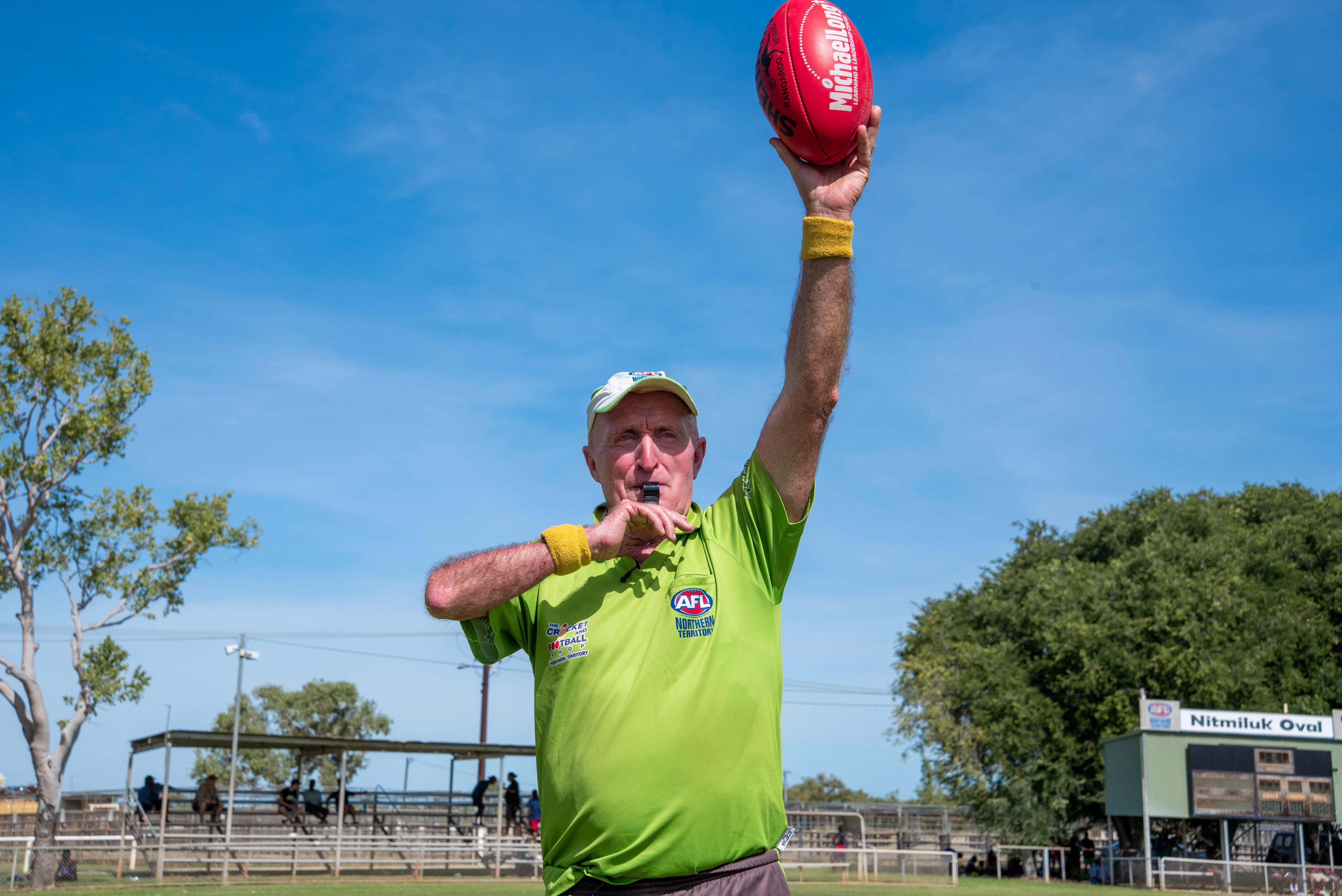 A man in a bright green t-shirt blows a whistle and holds a red AFL football in the air.