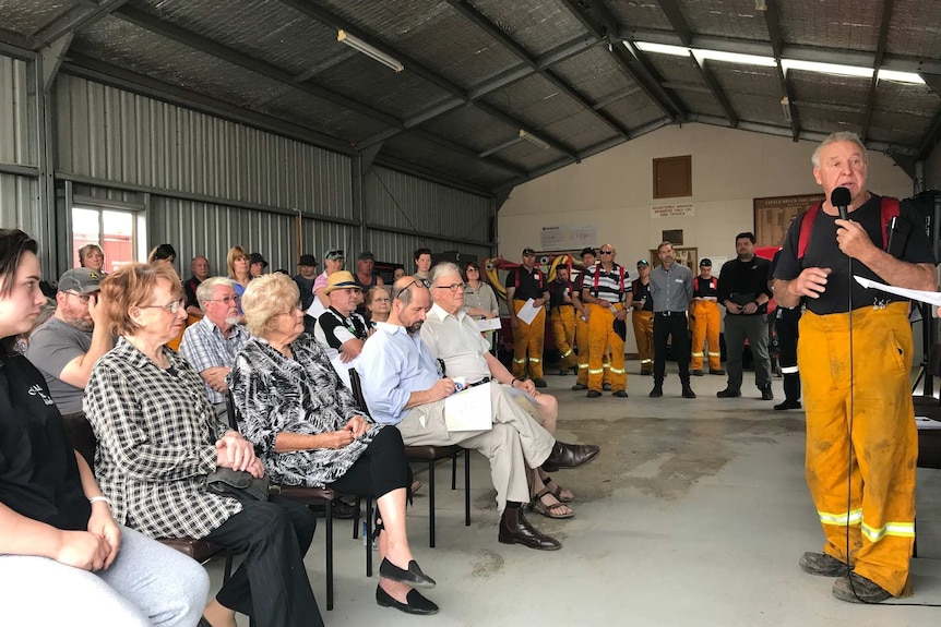 A group of men and women sit on plastic chairs in a tin shed as a man in overalls speaks into a microphone.