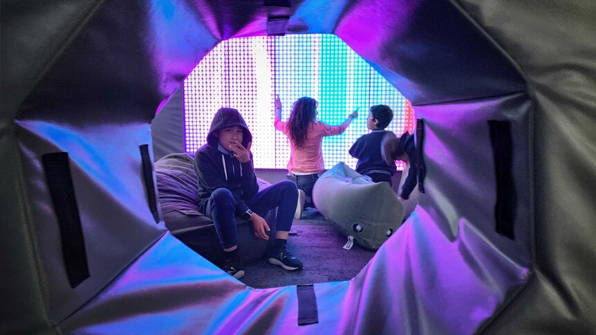 A picture of three students in a play area with a bright screen behind them and beanbags on the floo.