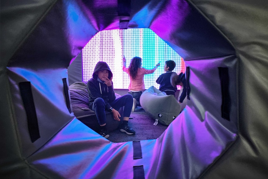 A picture of three students in a play area with a bright screen behind them and beanbags on the floo.