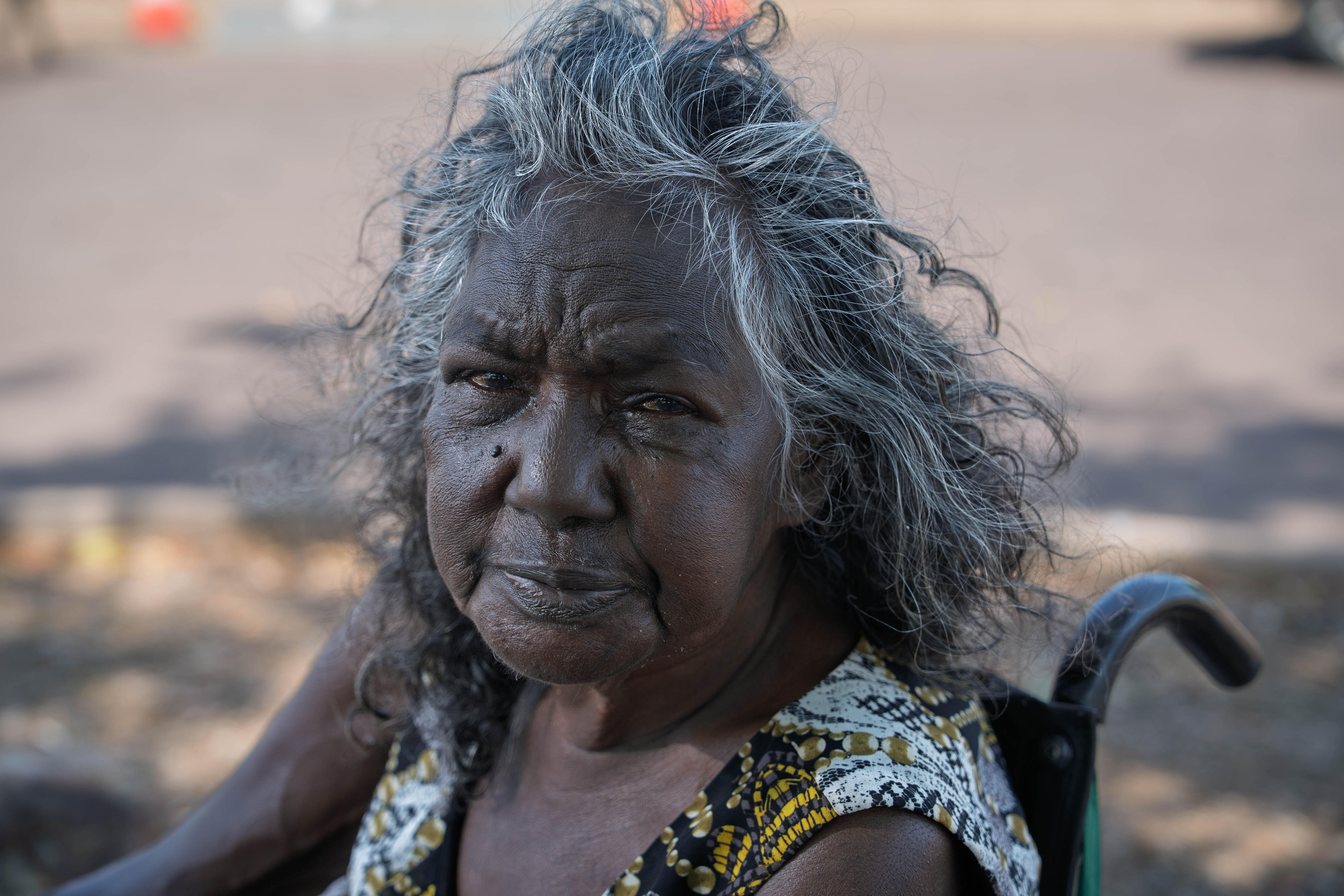 Evonne, an older Indigenous woman sitting in a wheelchair, looks up at the camera with a worried facial expression.