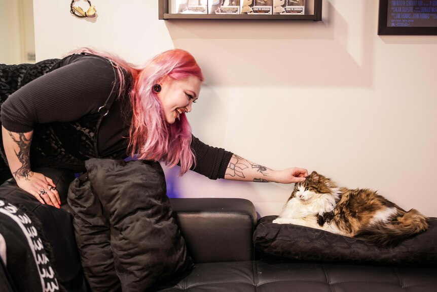 A woman smiles as she leans over a black couch to pat a fluffy cat. 