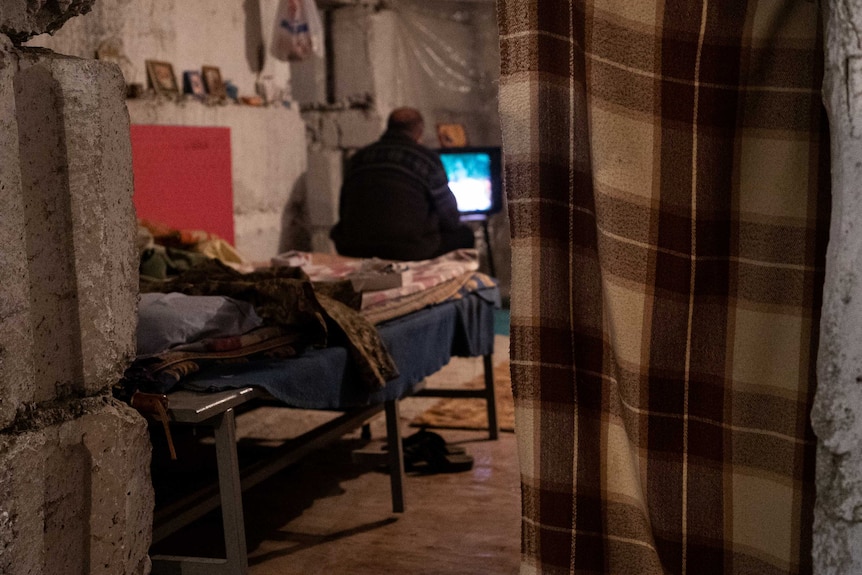 A man sits on a bed and watches TV in an underground room