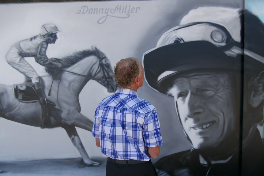 75-year-old jockey Danny Miller stands in front of a mural painted in his honour at the Bunbury Turf Club.