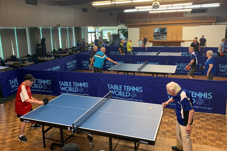 Two veteran table tennis players in a game at an indoor sports centre with other tables in the background