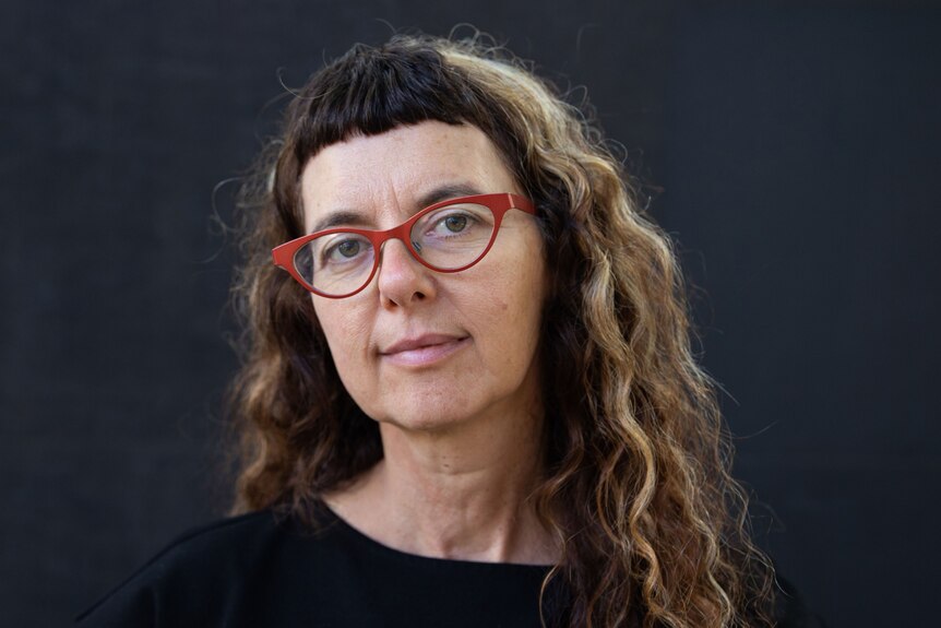A woman with long curly hair and red glasses looks into the camera with a half smile. 