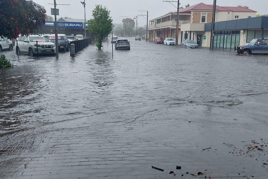 A flooded street with a car yard to the left and commercial buildings on the right and a few cars