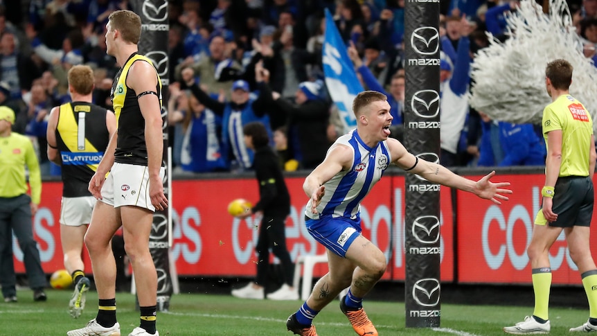 A North Melbourne AFL player runs away with his arms wide in celebration as Richmond players stand dejected in the background.