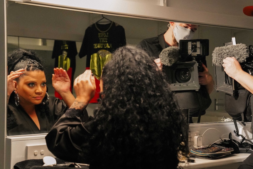 Singer-songwriter Naomi Wenitong prepares her hair and make-up in front of a mirror in a dressing room while being filmed.