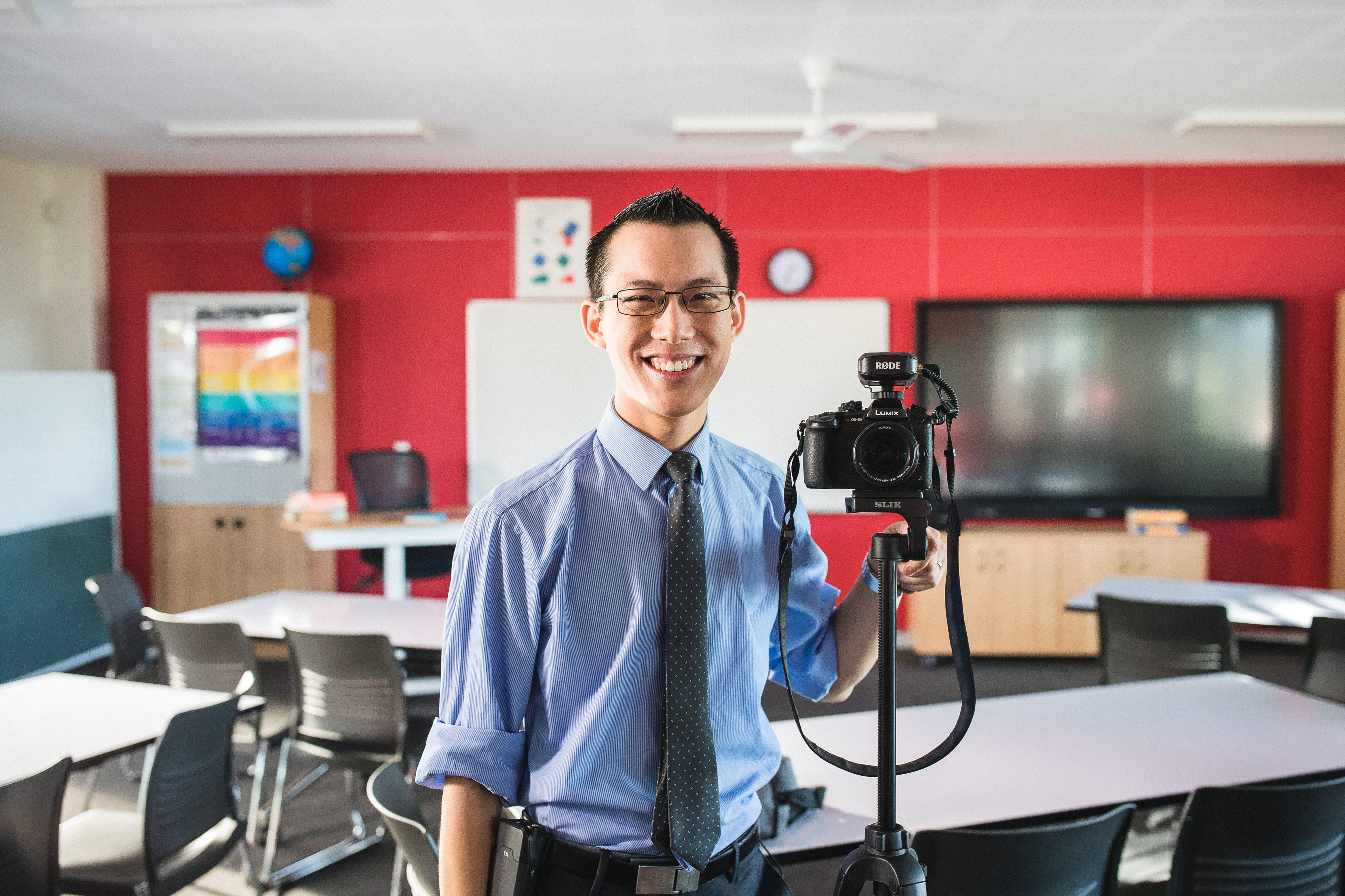 A smiling, dark-haired man in a shirt and tie stands next to a camera on a tripod in a classroom.