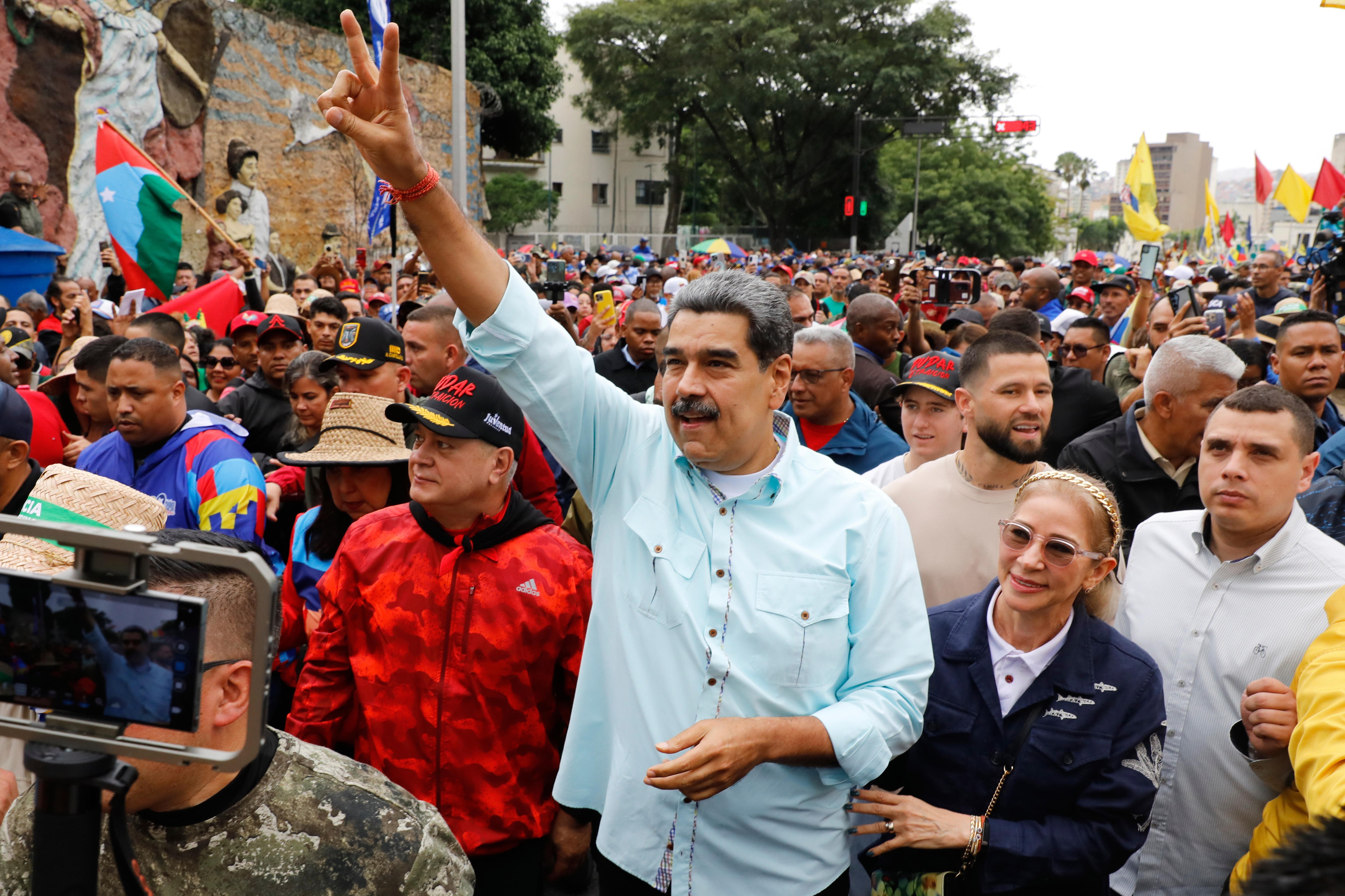 Nicolas Maduro in a light blue shirt raising a peace sign in a crowd of people at an outdoor rally