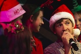 A man in a Christmas hat holds a microphone while singing on a dark stage surrounded by children.