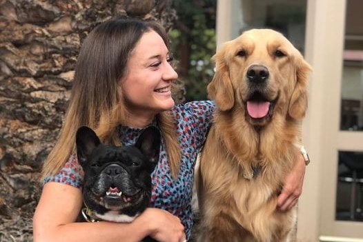 A smiling woman looks at a golden retriever with her arm around it. A small dog with black fur sits in her lap