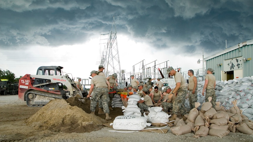American soldiers in light uniform help build a levee on a dirt patch in front of an electrical generator as dark clouds loom.