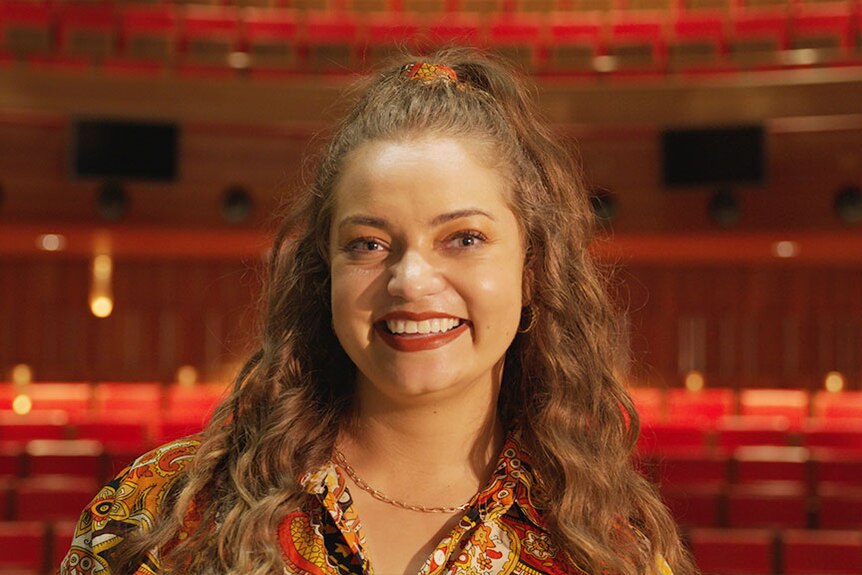 A young Aboriginal woman beams at the camera in a theatre.