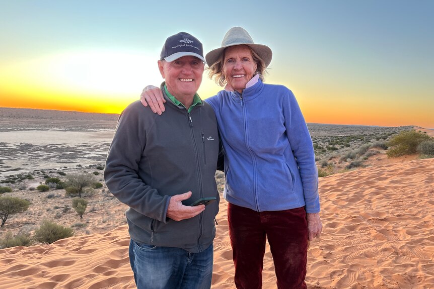 Man and woman stand arm in arm on a high sand dune with dramatic sunset in the background. 