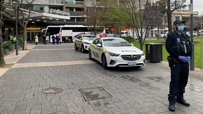 A police officer wearing a medical mask stands in front of two police cars with people and a bus in the background.