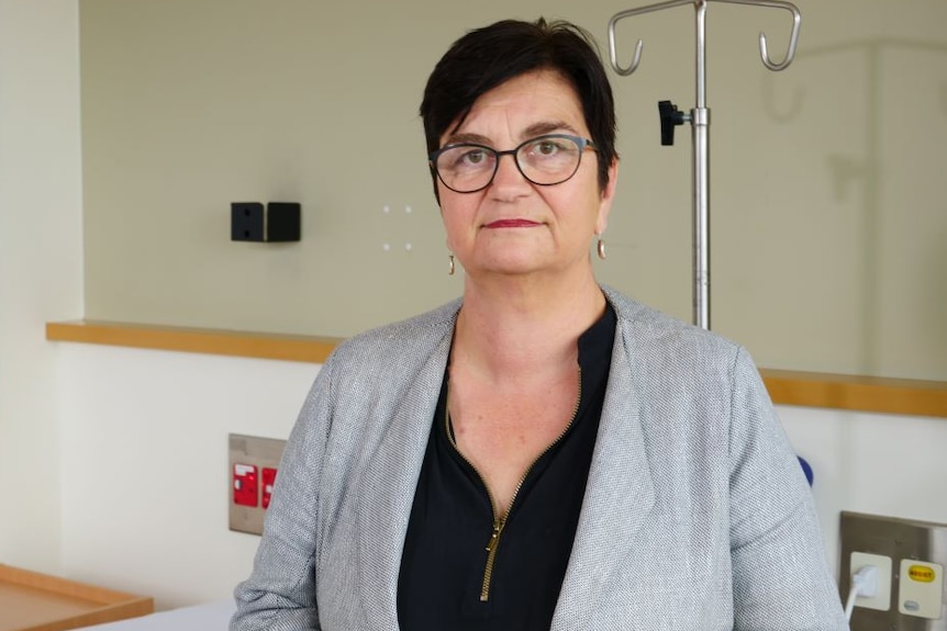 A woman in a black shirt and grey blazer stands by a hospital bed.