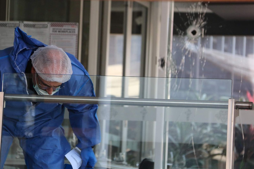 A forensic officer in a jump suit near a bullet hole visible in a window