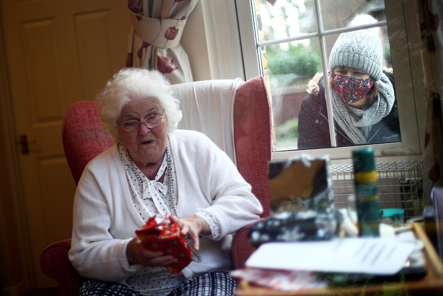 An older woman with white hair opens up a present while a masked woman looks through a window 
