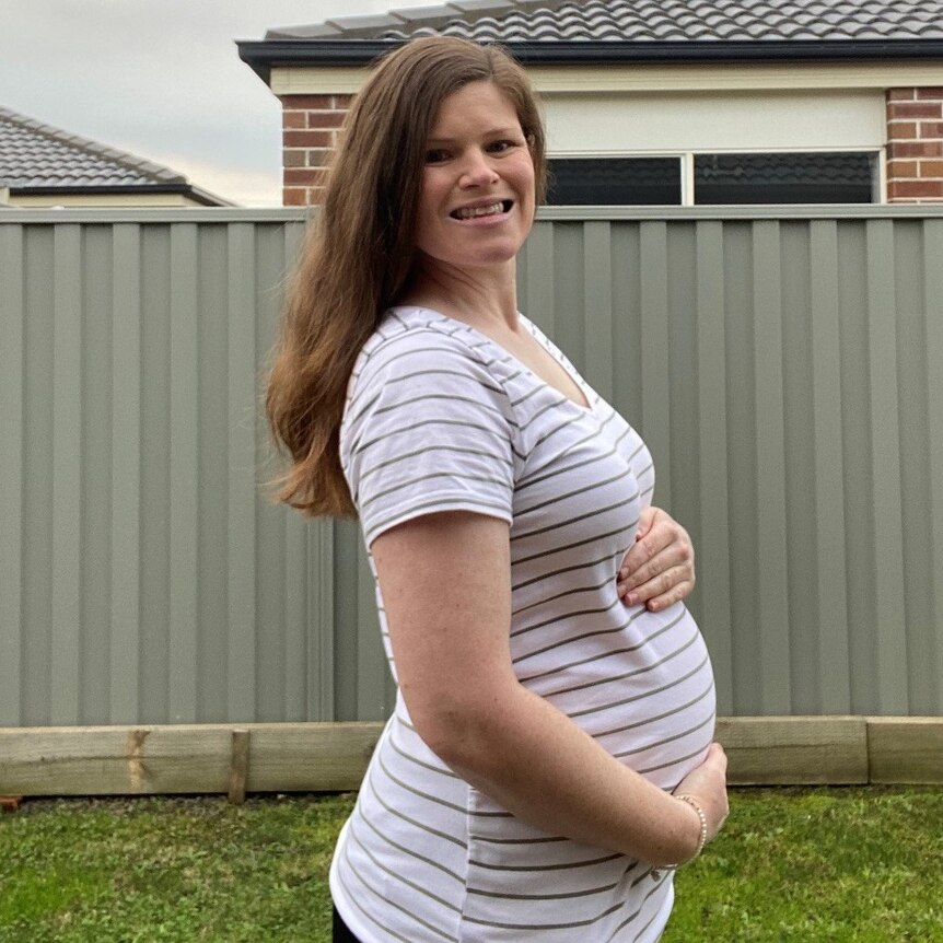 A lady rests her hand on her baby bump as she smiles at the camera while standing in her garden.