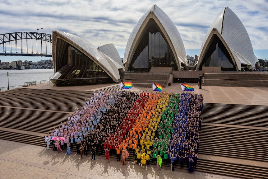 people in the formation of the rainbow flag standing on the steps of the opera house in sydney