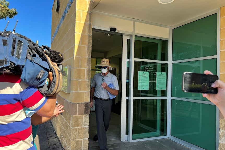 A man wearing a straw hat, sunglasses and a face mask exits a court building