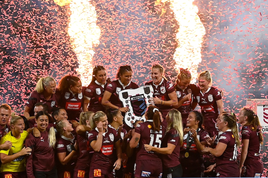 Queensland Maroons lift the Women's State of Origin shield as pyrotechnic flames erupt behind them.