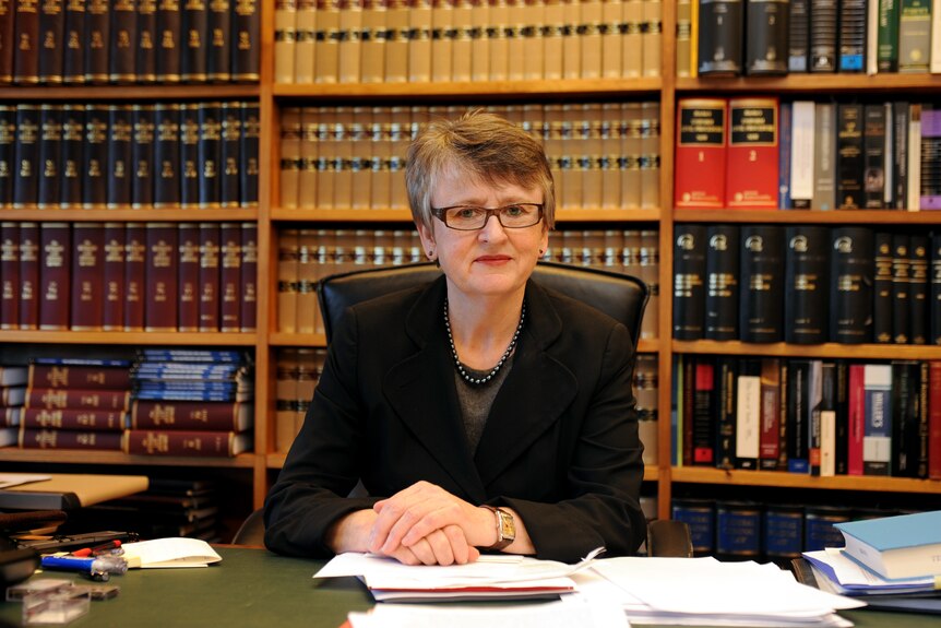 A woman with short hair and glasses sits at a desk in front of bookshelves lined with legal texts.