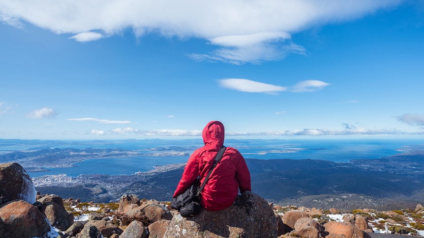 A person wearing a warm parka sits with their back facing the viewer, overlooking a stunning view of the land near Hobart.