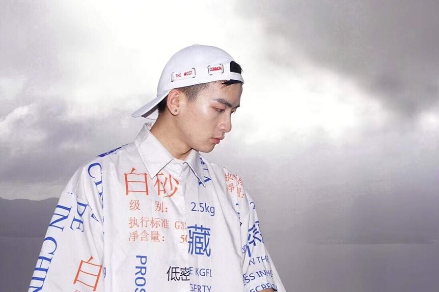 Zhang Yangzi poses in some of his fashionable white clothing covered in writing, standing in front of a cloudy sky