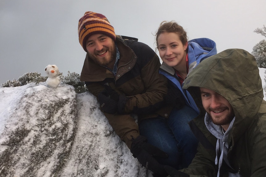 Three young people in snow gear pose next to a large snowy rock with a tiny snowman perched on top.