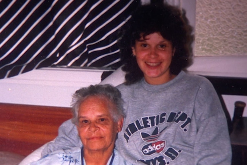 A woman wearing a grey sweatshjrt is seated with arm around her mother who has short grey hair