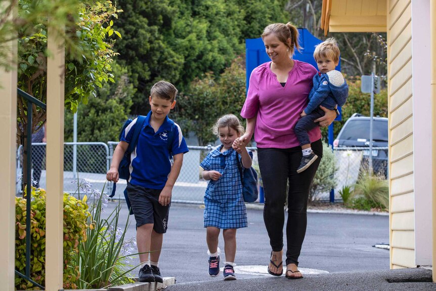 Woman carrying toddler walks with two primary school aged children dressed in uniform.