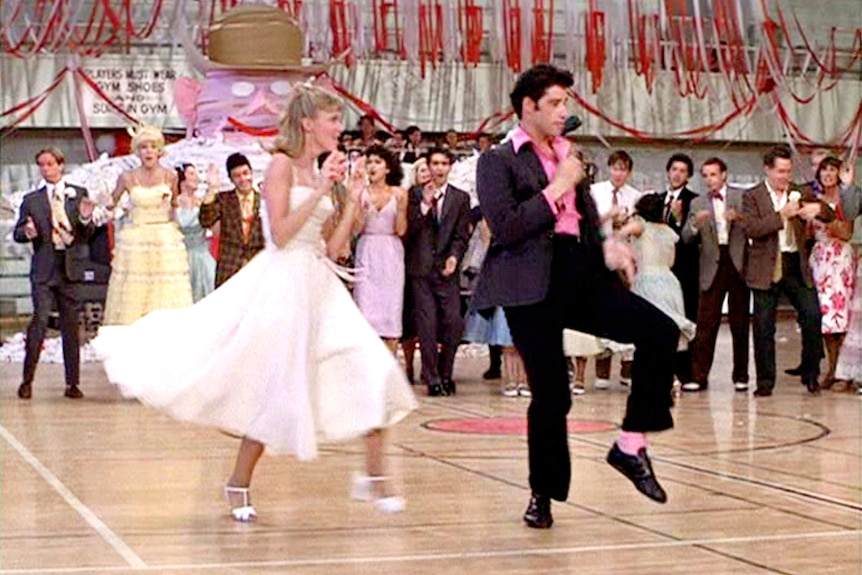Olivia Newton John and John Travolta dance in a decorated school hall surrounded by people. 